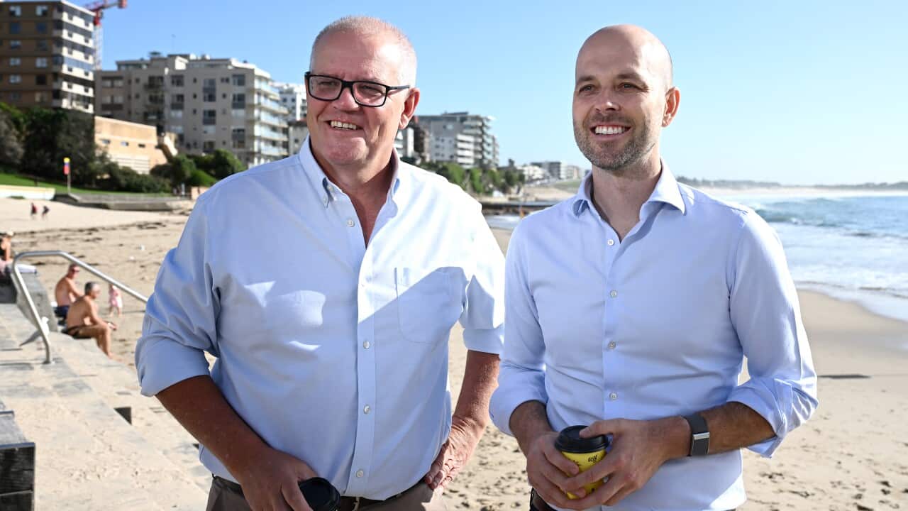 Two men stand smiling, with coffee cups in their hands against a beach backdrop.
