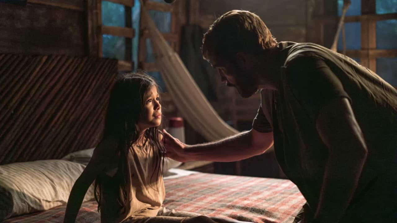 A young girl looking alarmed on a bed. A man leans in to comfort her by touching her shoulder.