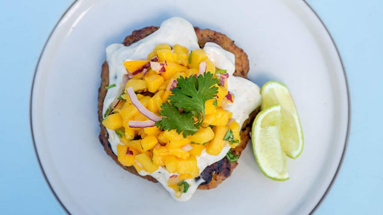 A brown patty sits on a wide white plate. The patty is topped with a creamy sauce. On top sits a chopped mango salsa.