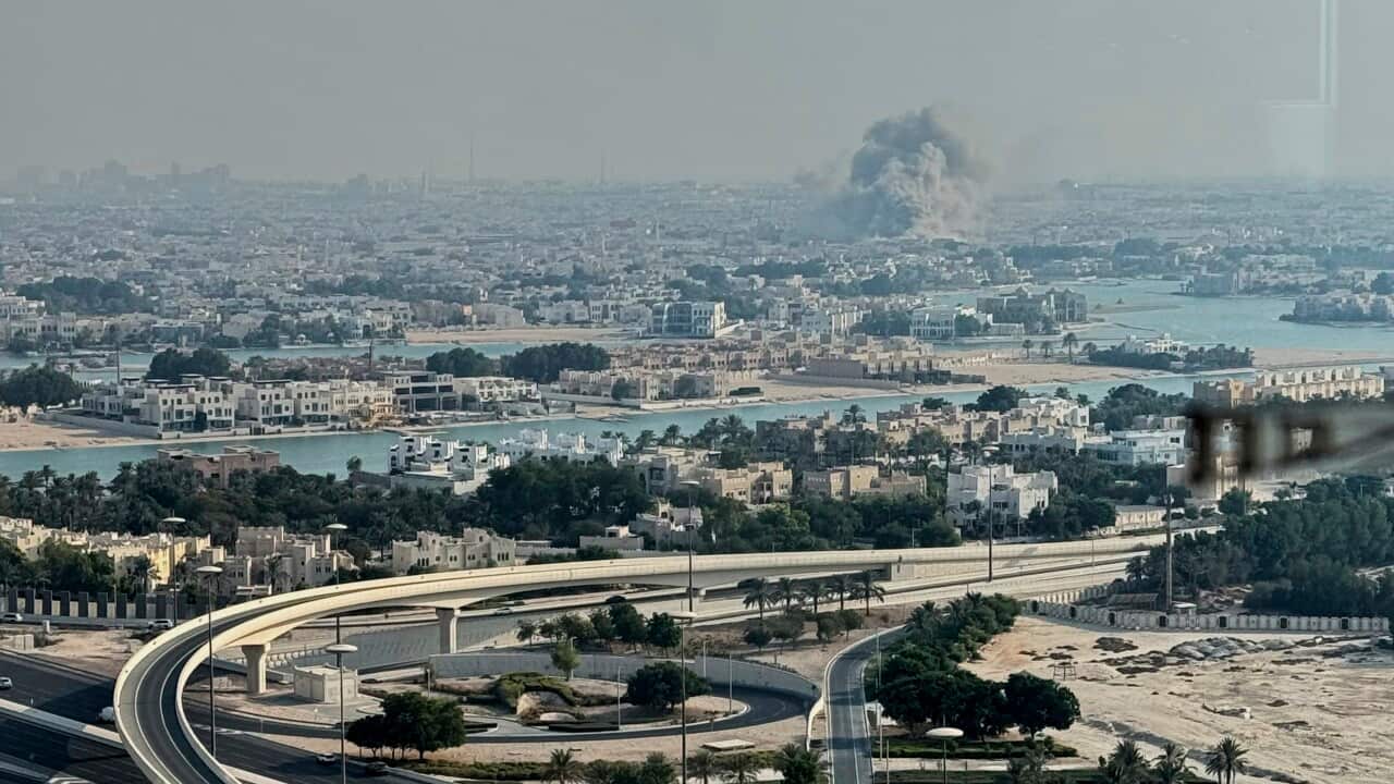 A plume of smoke rises from the site of an airstrike at a distance with a city in the foreground.