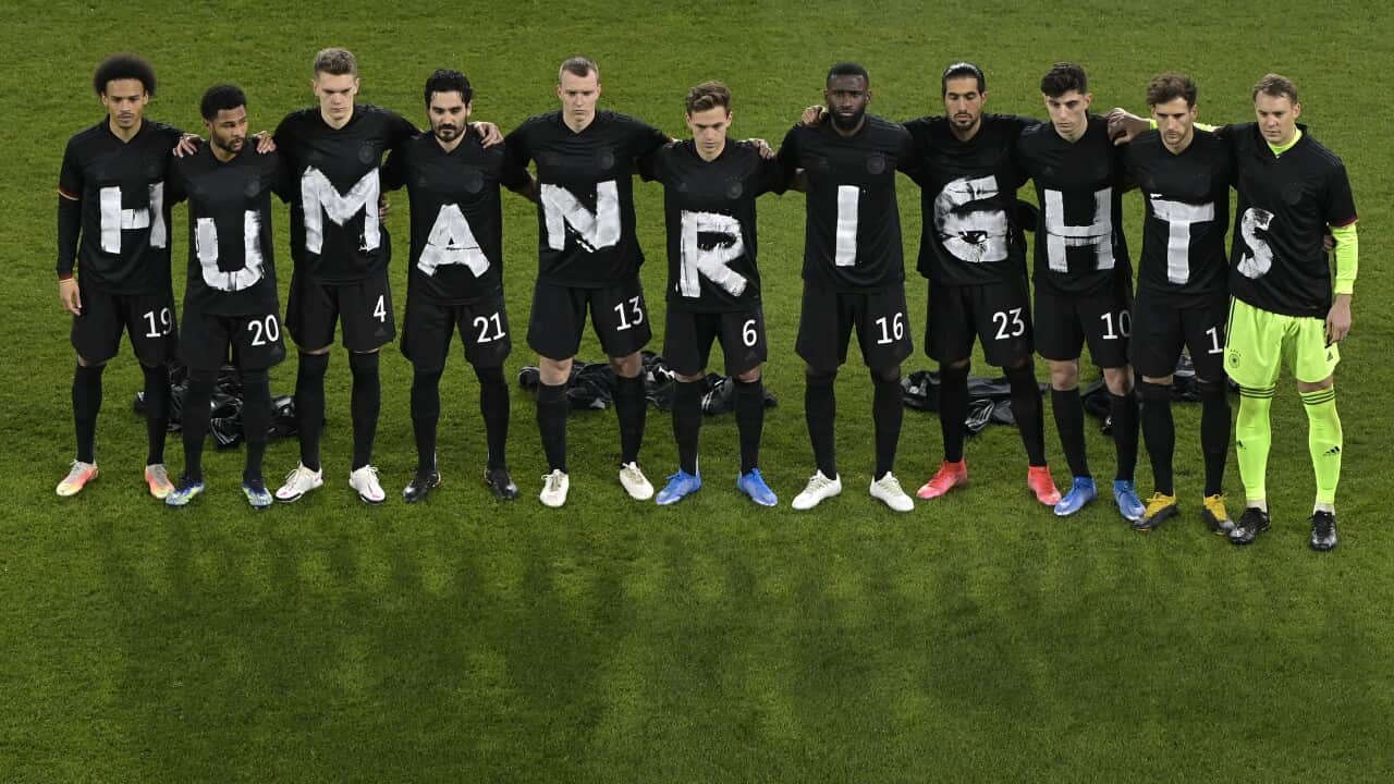 On a football pitch, a men's football team stand in a line. They are dressed in black T-shirts, with each shirt displaying a letter that together spells out the phrase "human rights".