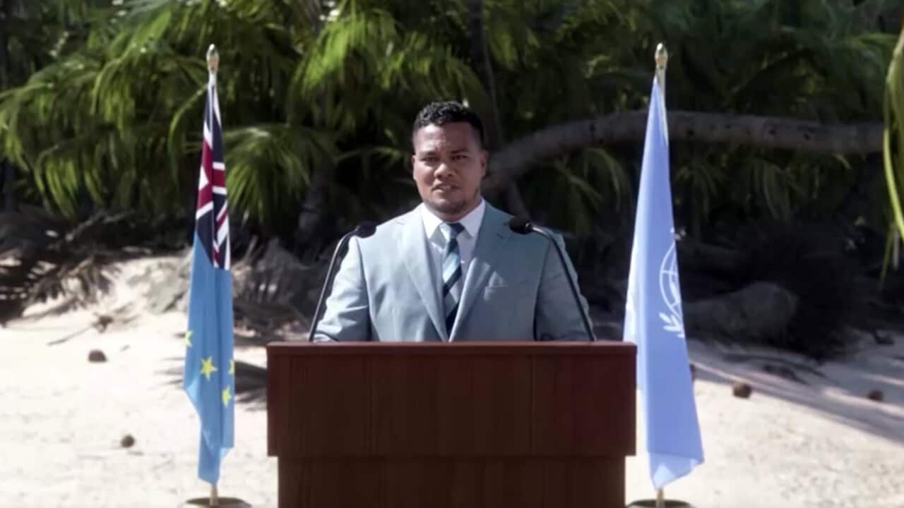 A man stands at a lectern on a beach with two flagpoles behind him