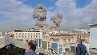 From a rooftop where men are standing, two large plumes of grey smoke rise high into the air over a densely populated city.