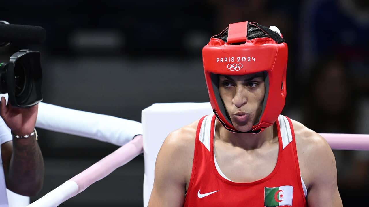 A woman in a red singlet and red boxing helmet stands with a determined expression on her face in the corner of a boxing ring.