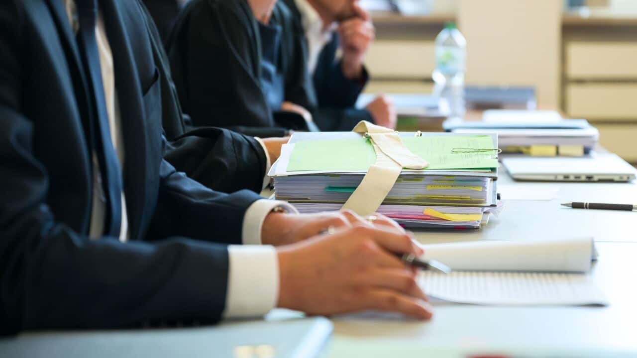 Three people sitting at a desk with large piles of documents