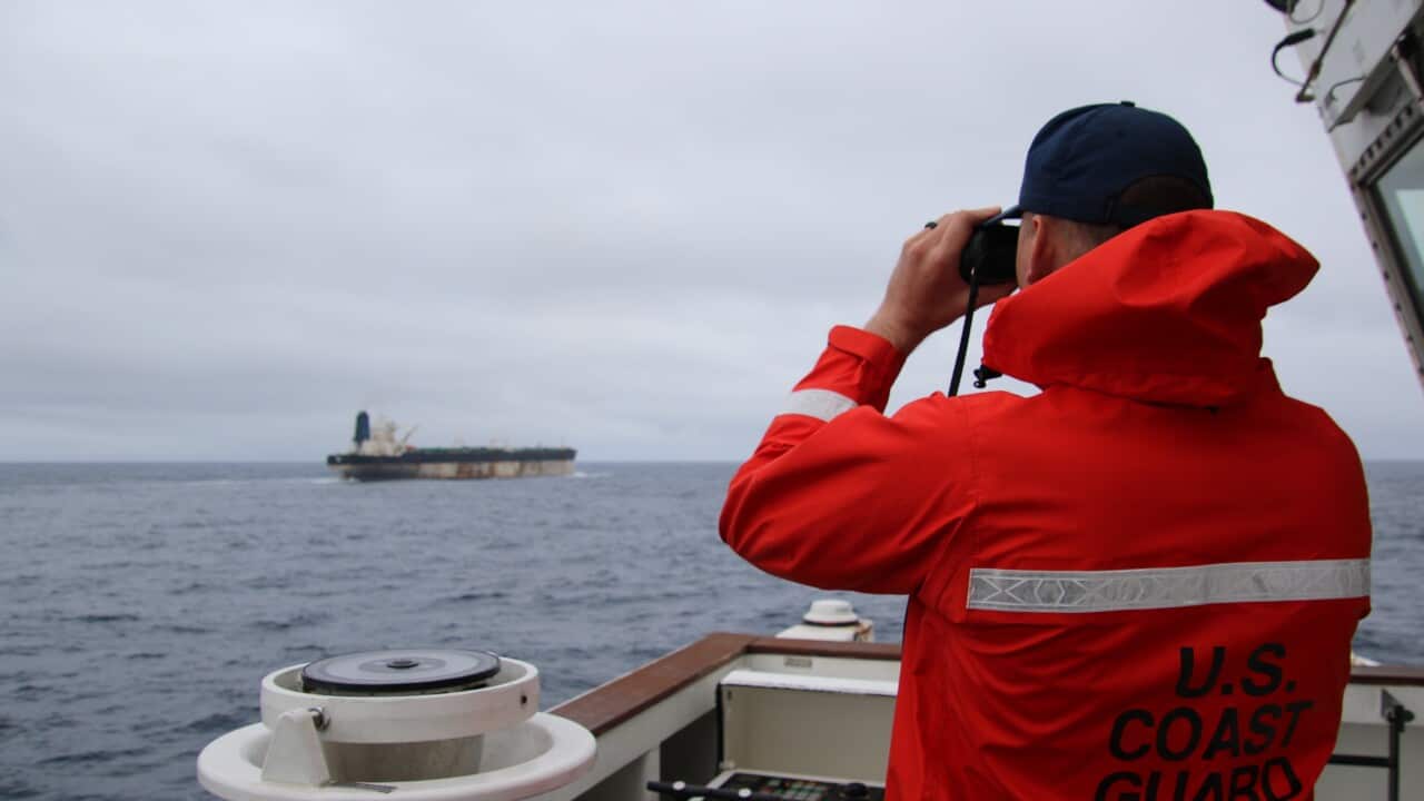 A person in a red US Coast Guard jacket peering through binoculars at an oil tanker in the distance.