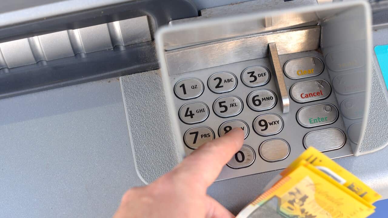 A close-up shot shows a person's finger pressing a button on an ATM keypad while they hold a fan of Australian dollar banknotes.