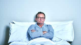 A man in striped pyjamas and glasses sits up in bed, leaning against the pillows. His arms are crossed and he has a wry, slightly glum smile. 
