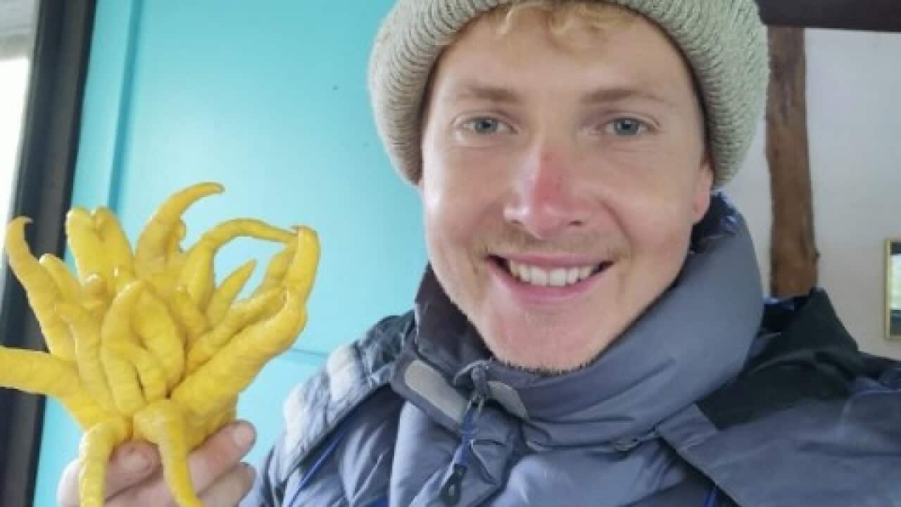 A young man in a grey beany and blue coat smiles at the camera and holds a twirly fruit in his hand.