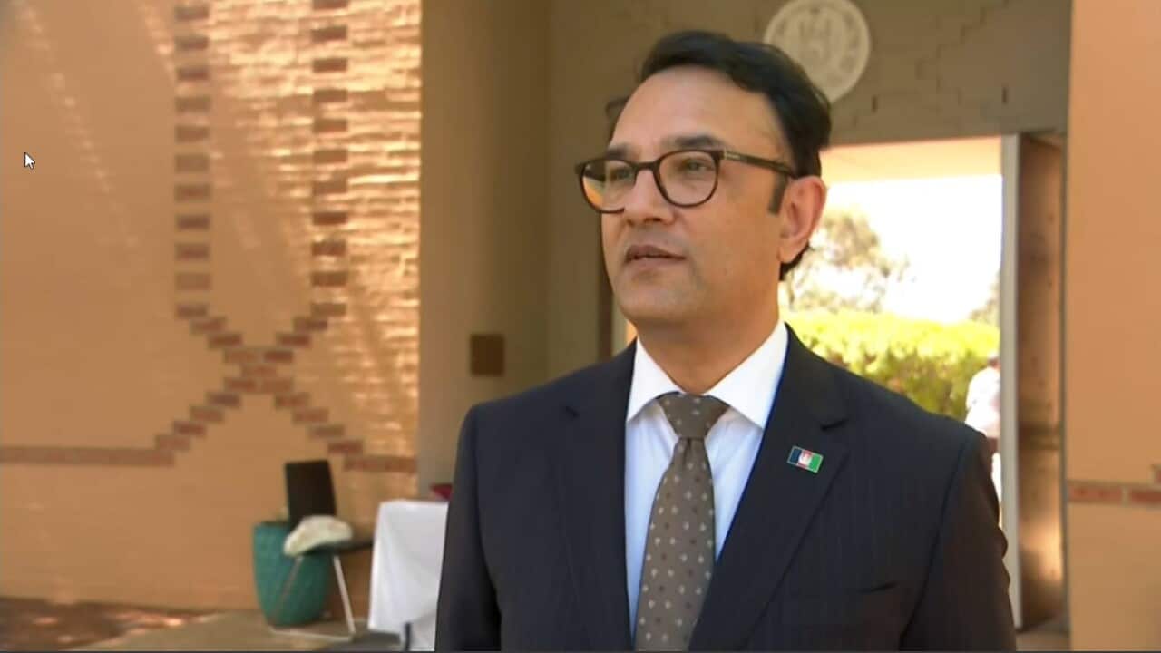 A man in a suit and glasses stands in front of an orange building.