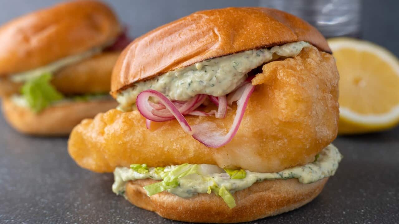 A thick golden battered fish fillet sits in a bud, with a green-speckled sauce and rings of pickled red onion. A second burger can be seen sitting behind.