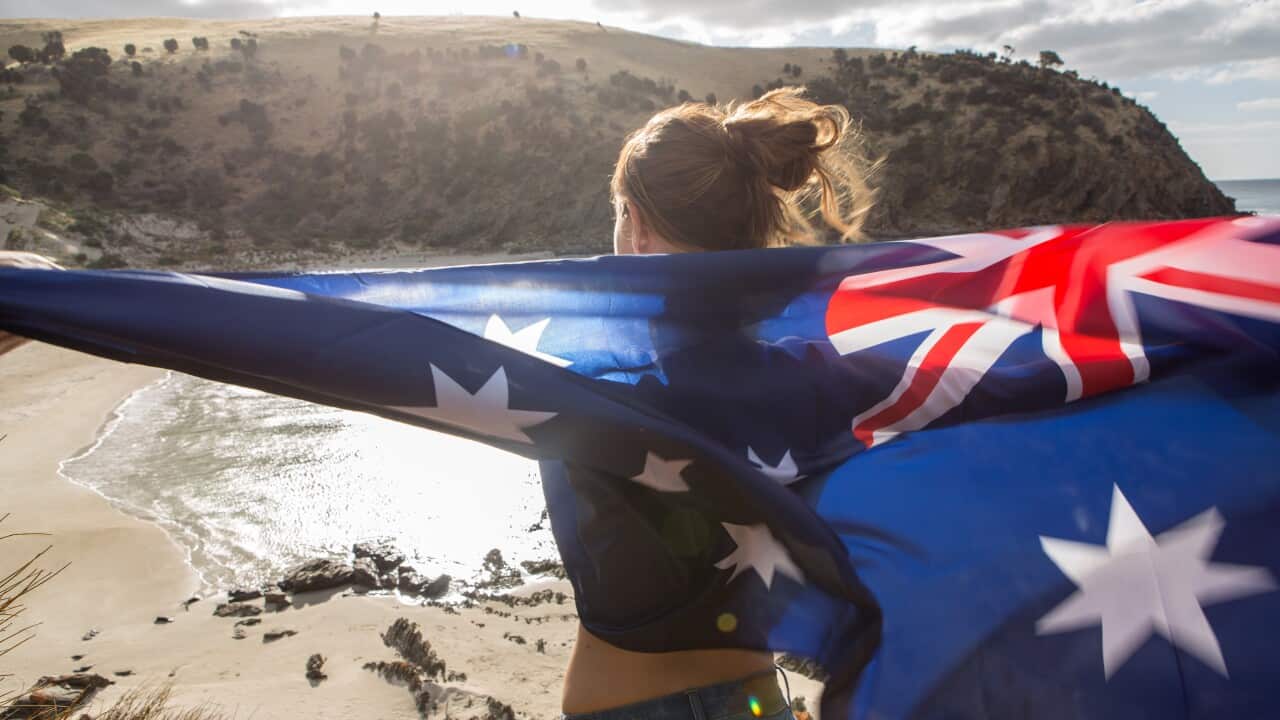 Girl stands on cliff above beach holding Australian's flag