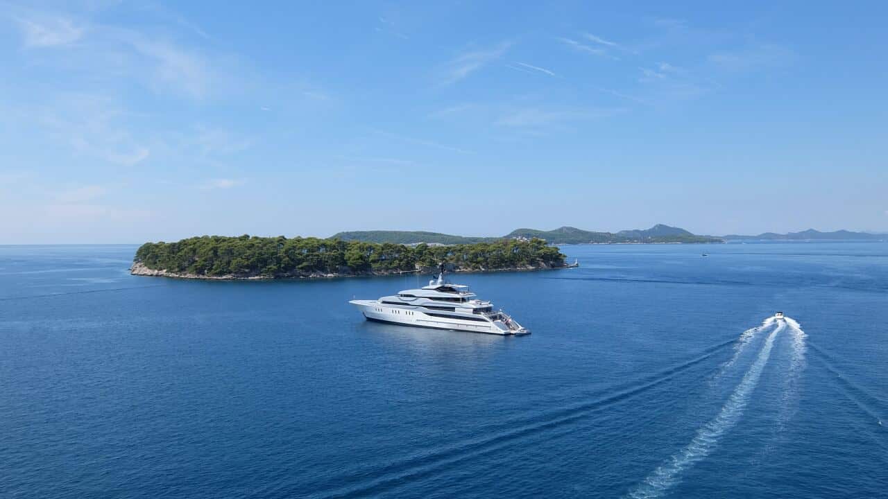 Landscape view of a verdant island surrounded by bright blue water, with a super yacht sailing across the midground