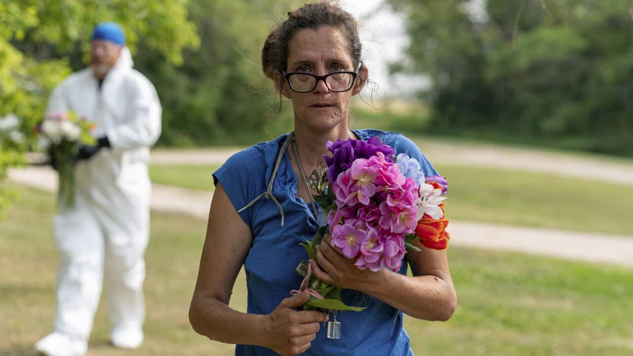A woman stands with a serious facial expression and carrying flowers.