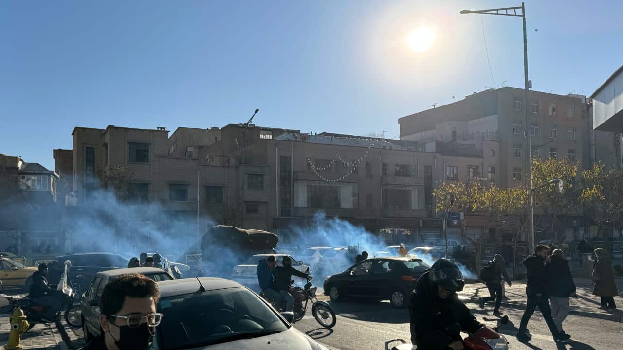 Smoke drifts through a busy street as people and motorbikes move between cars under a bright sun.