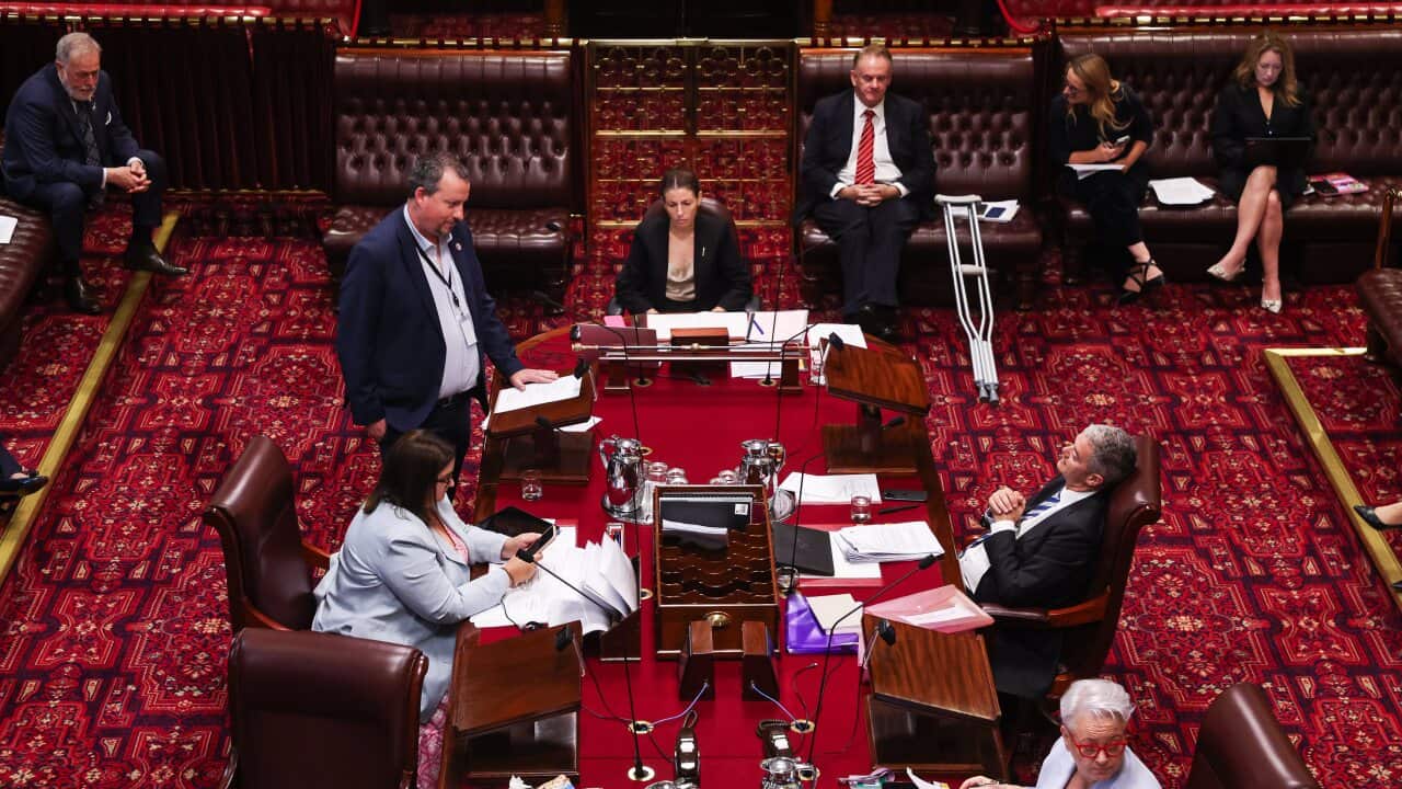 An overhead shot shows a group of people gathered around a long wooden table in a formal, red-carpeted chamber, likely a legislative assembly, with one man standing to address the others.