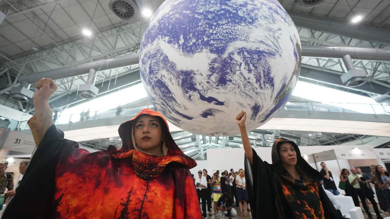 Two women in dark, hooded robes, one with a fiery red and black pattern and the other dark with an orange glow, stand indoors with their fists raised beneath a large, suspended inflatable globe.