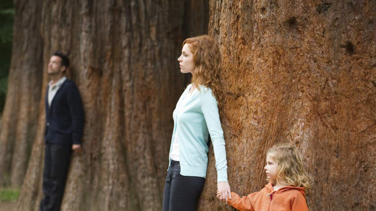 Mother and daughter leaning against tree, father standing separate in background