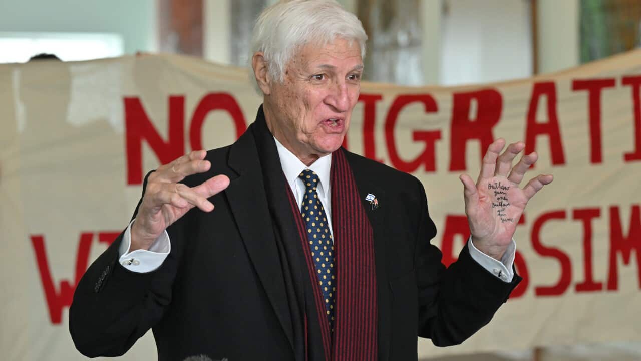 Bob Katter, an elderly man with white hair and wearing a black coat, gestures with his hands—one of which has writing on the palm—while standing in front of a banner reading “no migration without assimilation“.