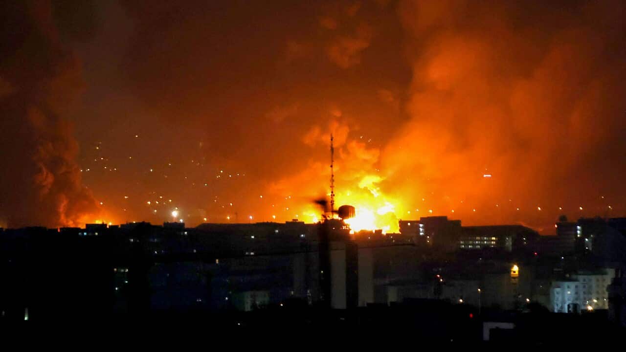 A massive orange fireball and thick smoke illuminate the night sky behind the dark silhouette of a city skyline.