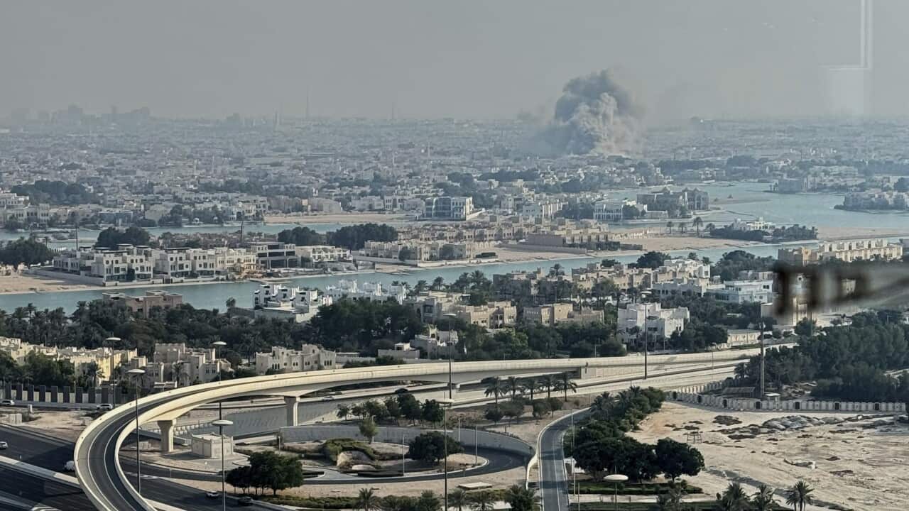 A plume of smoke rises from the site of an airstrike at a distance with a city in the foreground.