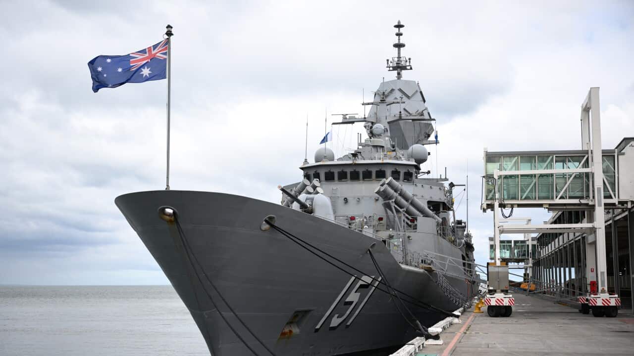 A large grey warship is docked at a pier with an Australian flag flying from its stern. The ship is viewed from the front, showing its a-frame mast and multiple missile launchers on its deck.