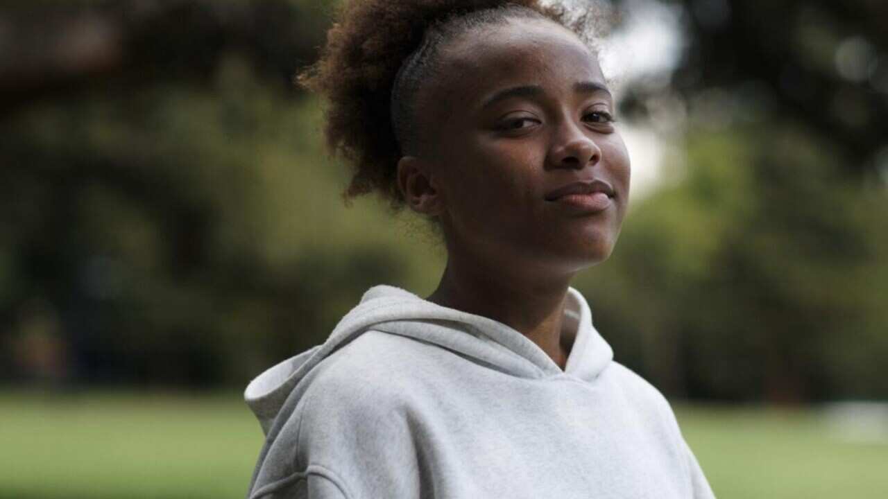 A young woman in a light grey jumper looks at the camera while at the park.