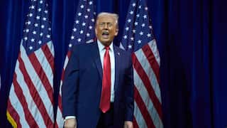 Donald Trump stands in front of three American flags and a blue curtain, appearing to speak or shout with an expressive facial gesture.