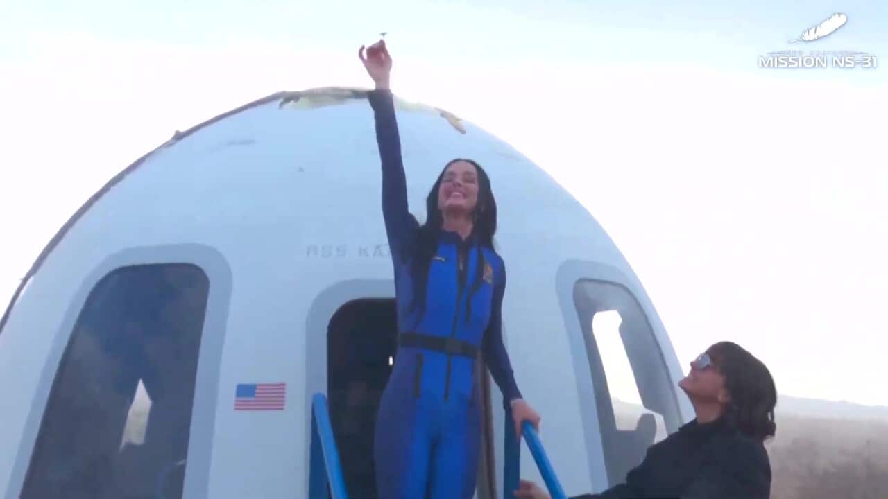 Katy Perry, wearing a blue space suit, holds a small flower high above her head as she stands on steps outside a white capsule. A woman to her right is looking up at her.