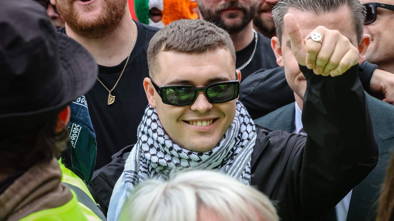 A young man with short hair and sunglasses, wearing a black jacket and a black and white keffiyeh, smiles directly at the camera while surrounded by a crowd. In the background, another man in a suit gives a thumbs-up sign, and a person is visible wearing a mask decorated with the Irish tricolor flag.