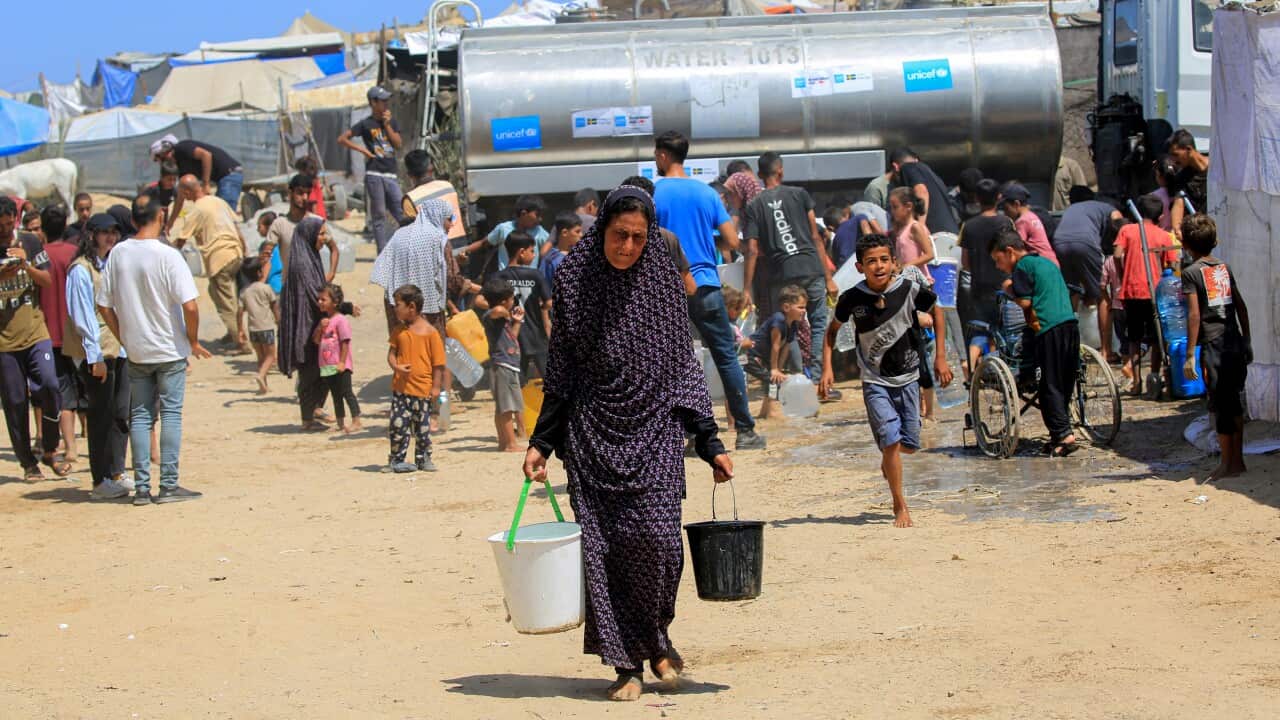 A woman walks with two buckets of water, a tanker, and several people, including children, in the background.