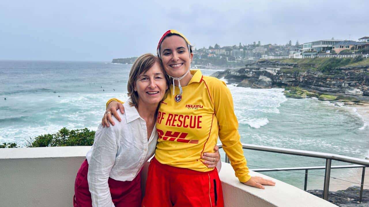 Sandra Fox, in white linen shirt and pink trousers is embraced by Ce Benedict in red and yellow life saver patrol uniform. The beach and coastline is behind them.