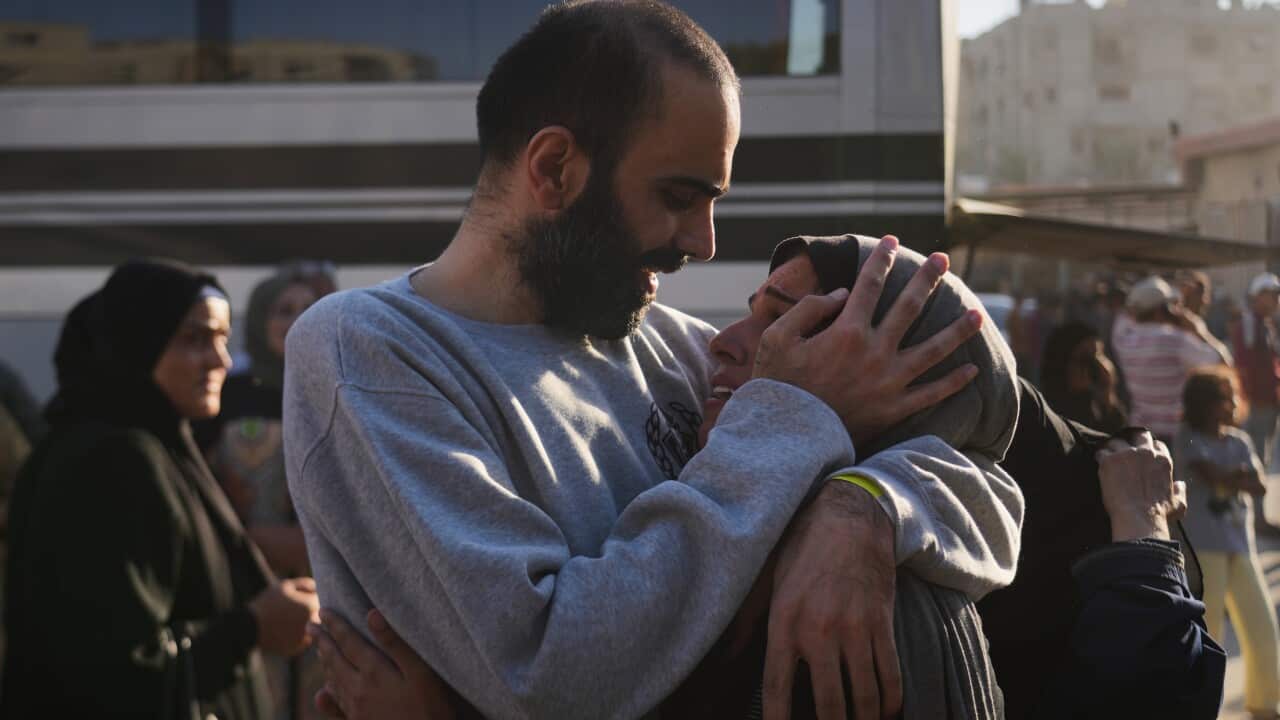 A man in a grey sweatshirt emotionally embraces a woman in a hijab, placing his hand against the side of her head and looking into her eyes. A bus and other people are in the backdrop.