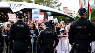 Three police officers with "New South Wales Police" patches on their vests stand with their backs to the camera, monitoring a crowd of protesters holding signs and flags at an outdoor rally.