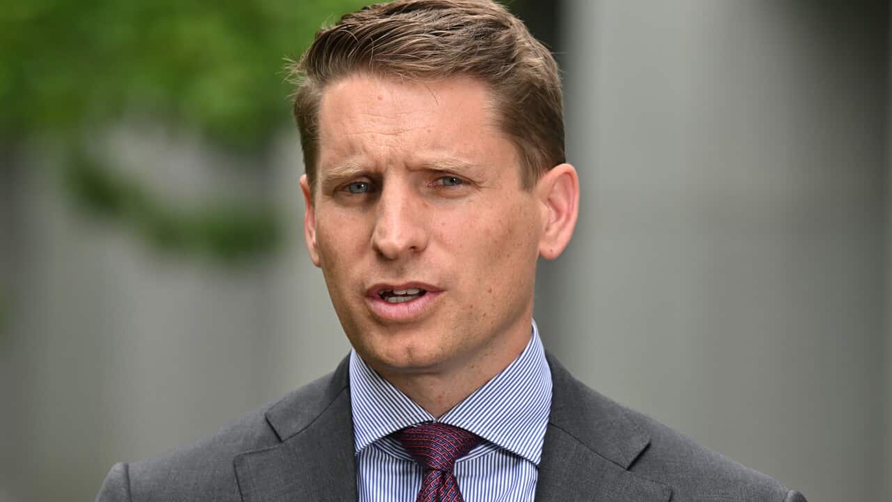 A close-up photograph features a man in a grey suit, striped blue shirt, and a patterned red tie, speaking outdoors with a serious expression. He has short, styled brown hair and appears to be a politician or public figure delivering a statement.