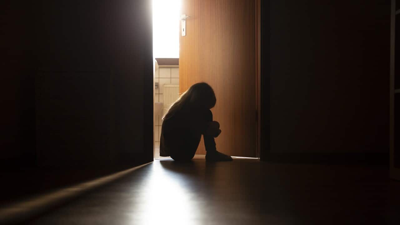 Despairing child sitting with head on knees in the dark frame of a doorway, backlit by a room behind flooded with daylight