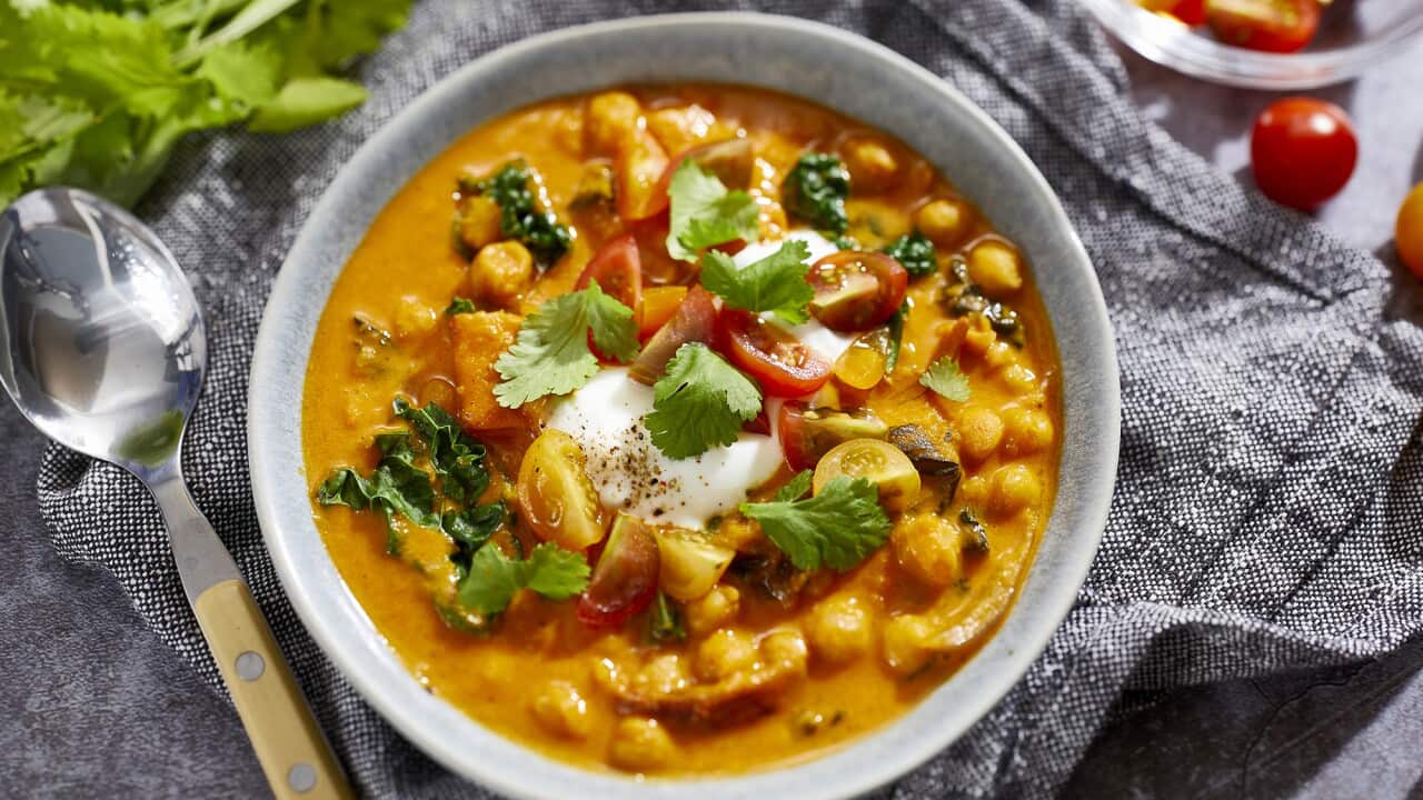 A pale bowl sits on a grey-toned cloth napkin. It holds a vibrant orange-yellow soupy stew, studded with chickpeas and vegetables. A metal soup spoon with a cream handle sits alongside.