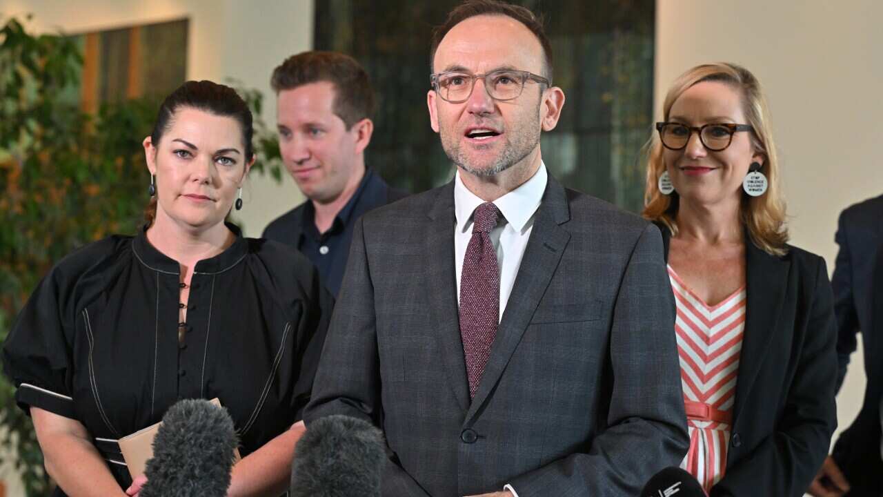 A man in a dark grey tweed suit speaks, looking ahead of him with two women on either side.