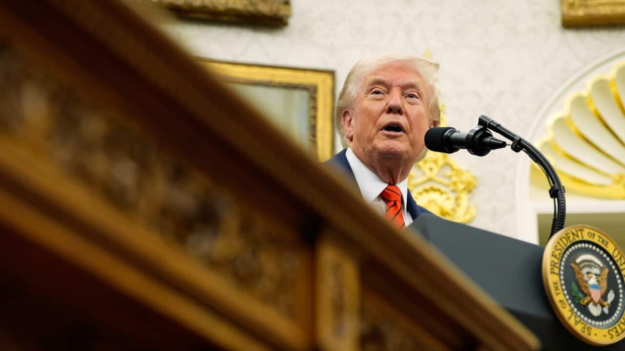 A blurred image of the rim of a golden carving of a table in the foreground, with Donald Trump speaking at a lectern in the background.