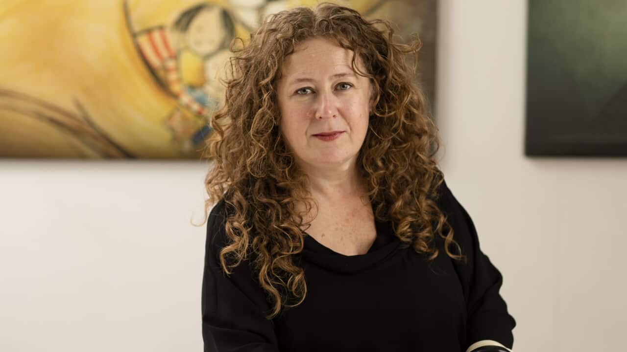 A woman with long curly brown hair rests her arms on a table.
