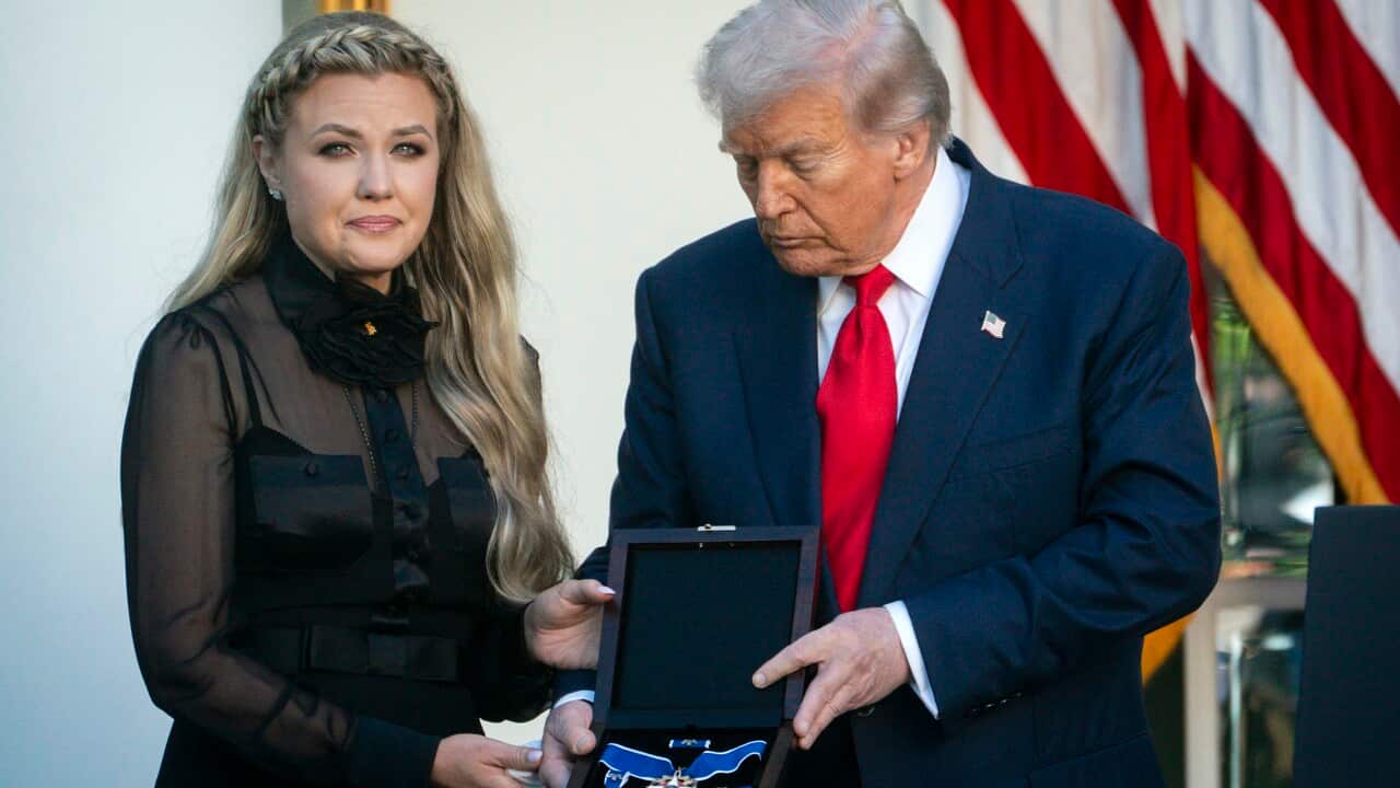 A woman in an all black outfit stands next to Donald Trump as he presents a medal to her with the American flag in the background.