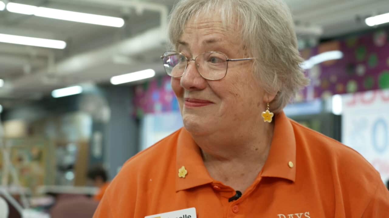 An elderly woman with short grey hair and glasses smiles warmly while wearing an orange polo shirt adorned with yellow flower pins.