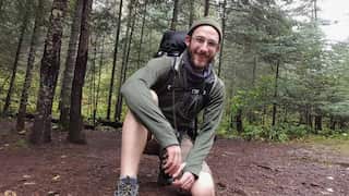 A white man wearing outdoor gear and a backpack kneels in some woods.