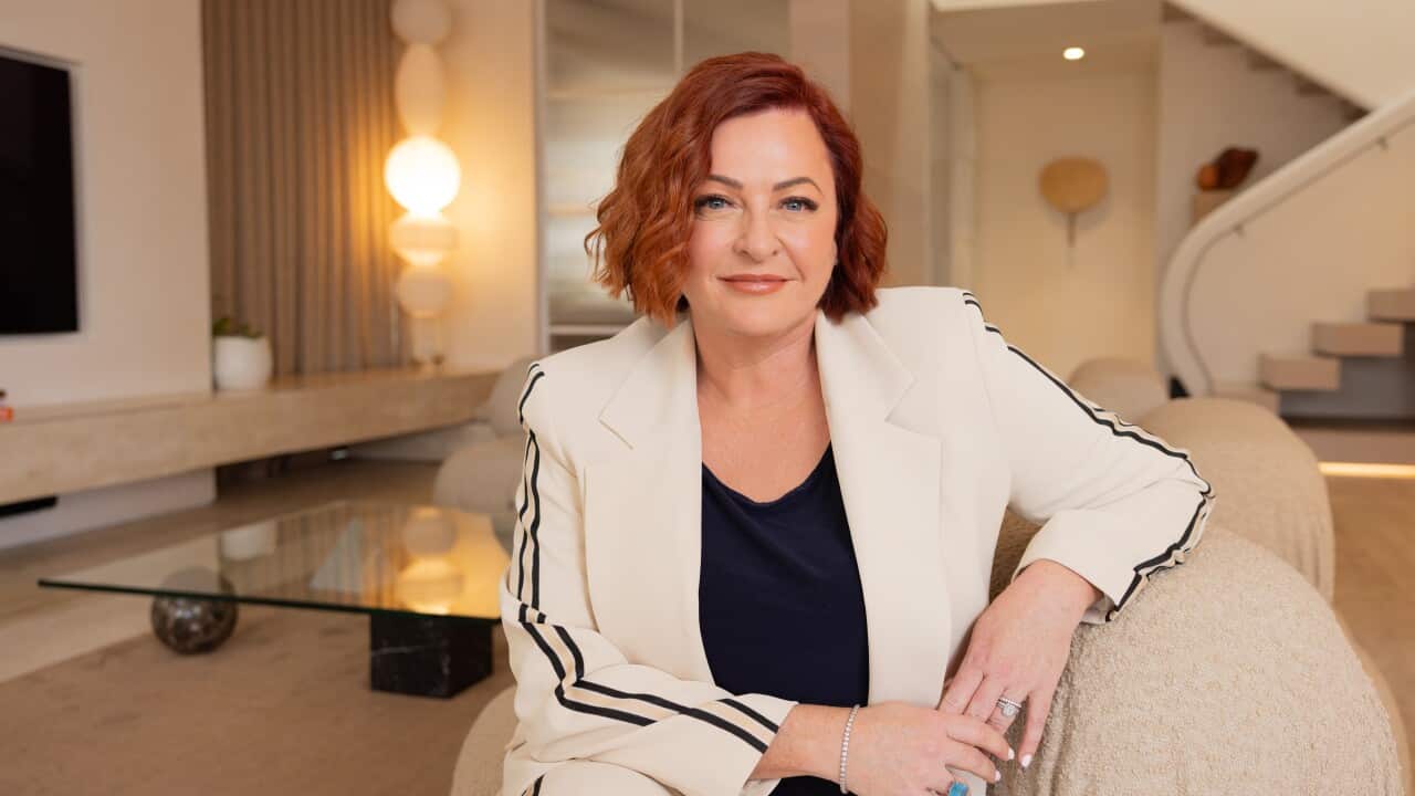 A woman in formal dress sits on an armchair in a luxurious loungeroom and looks at the camera with a neutral expression.