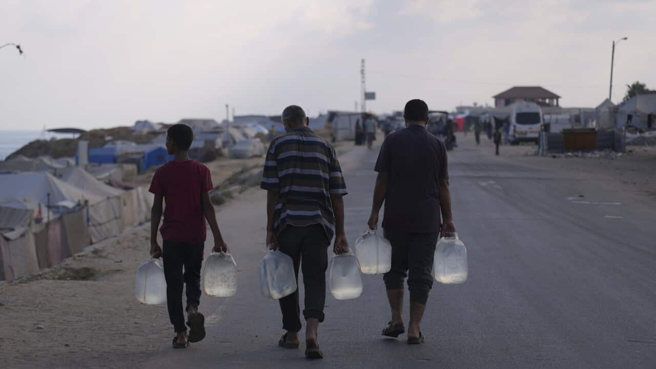 A photo taken from behind shows Palestinians carry plastic bottles with water near rows of tents.