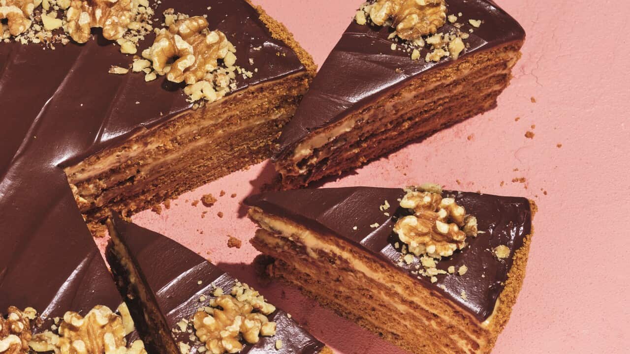 A round cake topped with chocolate icing and walnut halves sits on a peachy-red surface. Three slices have been cut from the cake and sit slightly away from it, showing the cake layers within.
