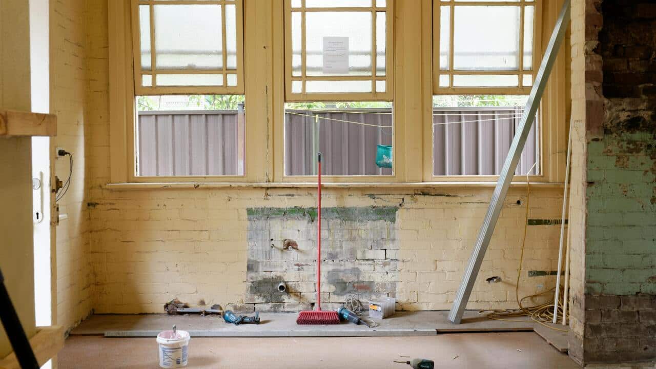 An unfinished wall in a house with a red broom standing in front of it. A bucket and tools are scattered on the floor