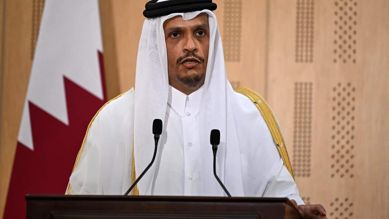 Un hombre con túnica habla en un podio frente a una bandera de Qatar.