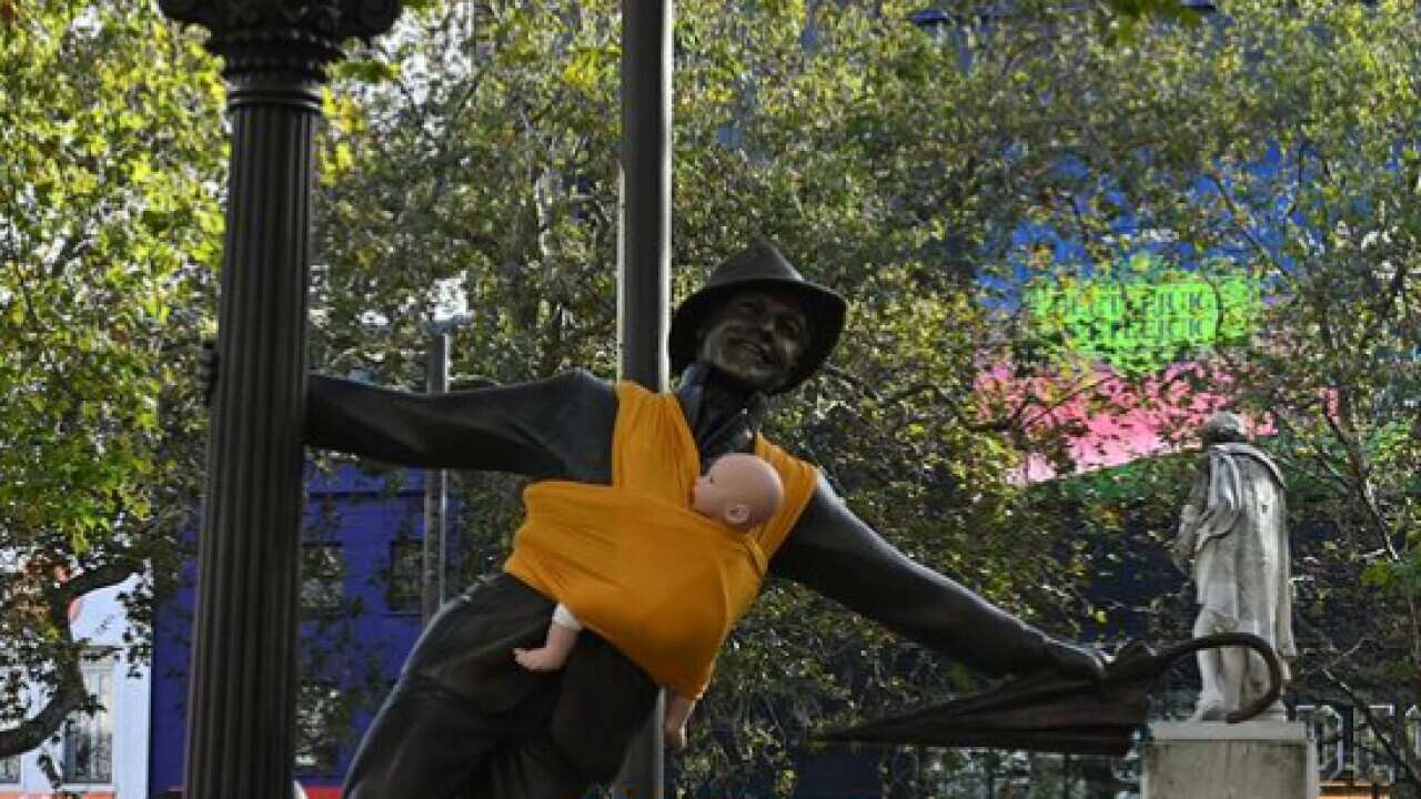 A statue of a man hanging from a lamppost, holding an umbrella and with a baby doll strapped to his chest using a yellow sling.