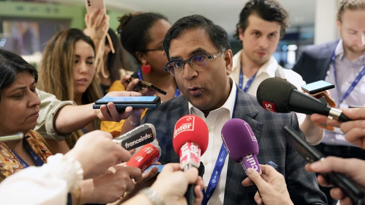 Avinash Persaud is surrounded by reporters holding microphones during a press conference.